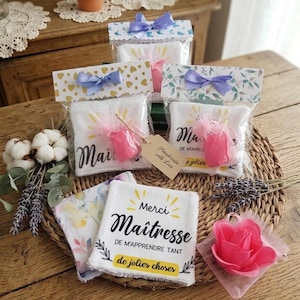 Könnte beinhalten: Mehrere kleine Geschenkbeutel mit weißen Handtüchern, jeweils mit einer Schleife gebunden. Einige Beutel haben bedruckte Oberteile mit goldenen Herzen oder floralen Mustern. Die Handtücher haben französischen Text und sind mit rosa Tüll und einer rosa Blume verziert.