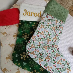 K&ouml;nnte beinhalten: Drei Weihnachtsstr&uuml;mpfe mit verschiedenen Mustern. Ein Strumpf ist gr&uuml;n mit goldenen Schneeflocken, einer ist wei&szlig; mit einem gr&uuml;nen, roten und goldenen Tannenzapfenmuster und einer ist beige mit goldenen Rentieren. Der gr&uuml;ne Strumpf hat den Namen "Jarod" in Gold gestickt.