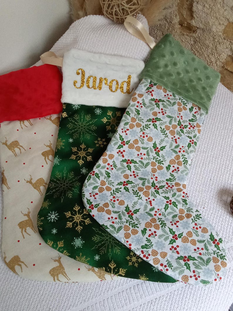 K&ouml;nnte beinhalten: Drei Weihnachtsstr&uuml;mpfe mit verschiedenen Mustern. Ein Strumpf ist gr&uuml;n mit goldenen Schneeflocken, einer ist wei&szlig; mit einem gr&uuml;nen, roten und goldenen Tannenzapfenmuster und einer ist beige mit goldenen Rentieren. Der gr&uuml;ne Strumpf hat den Namen "Jarod" in Gold gestickt.