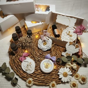 May include: A collection of white gift boxes, essential oil bottles, and decorative ornaments arranged on a woven mat. The ornaments include a bee, a rose, and an angel design. Dried flowers and daisies add to the aesthetic.