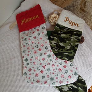 K&ouml;nnte beinhalten: Zwei Weihnachtsstr&uuml;mpfe, einer rot mit wei&szlig;en Schneeflocken und dem Wort "Maman" in Gold gestickt, und einer wei&szlig; mit Tarnmuster und dem Wort "Papa" in Gold gestickt.