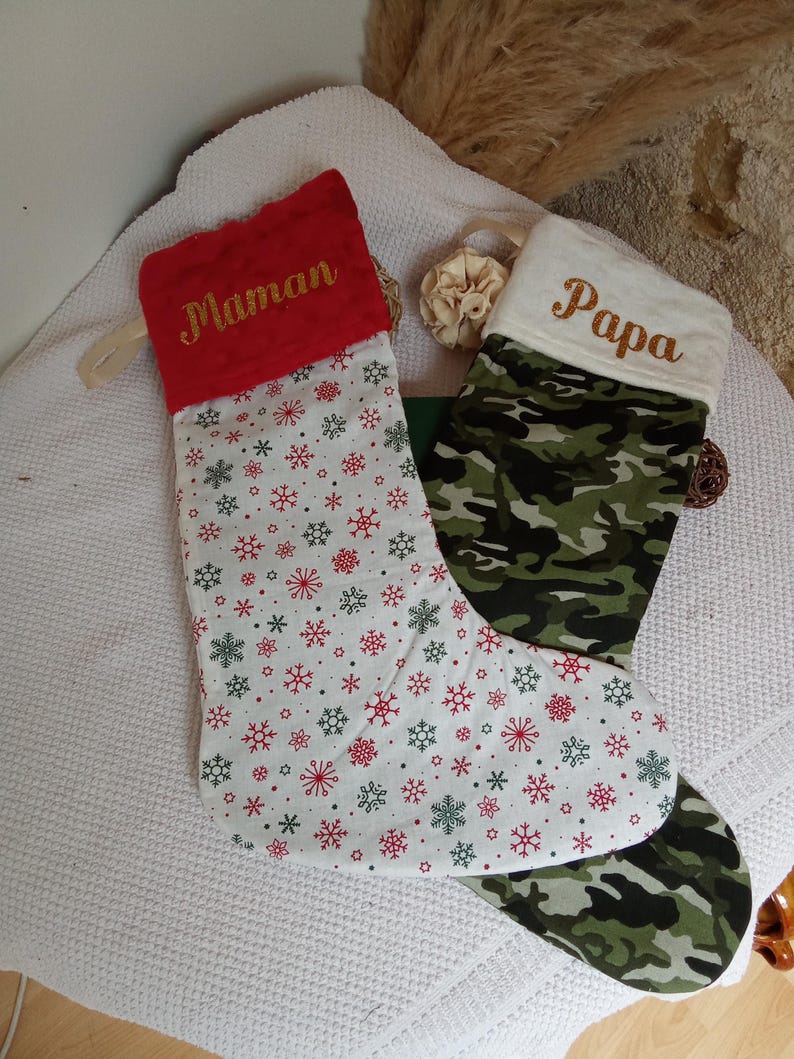 K&ouml;nnte beinhalten: Zwei Weihnachtsstr&uuml;mpfe, einer rot mit wei&szlig;en Schneeflocken und dem Wort "Maman" in Gold gestickt, und einer wei&szlig; mit Tarnmuster und dem Wort "Papa" in Gold gestickt.