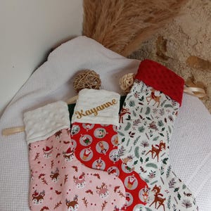 K&ouml;nnte beinhalten: Drei Weihnachtsstr&uuml;mpfe mit einem Rentiermotiv. Der Strumpf links ist rosa mit wei&szlig;en Punkten, der mittlere Strumpf ist rot mit einem Weihnachtsmannmotiv und der rechte Strumpf ist wei&szlig; mit einem gr&uuml;nen und braunen Rentiermotiv. Der mittlere Strumpf tr&auml;gt den Namen "Kayanne" in goldener Stickerei.