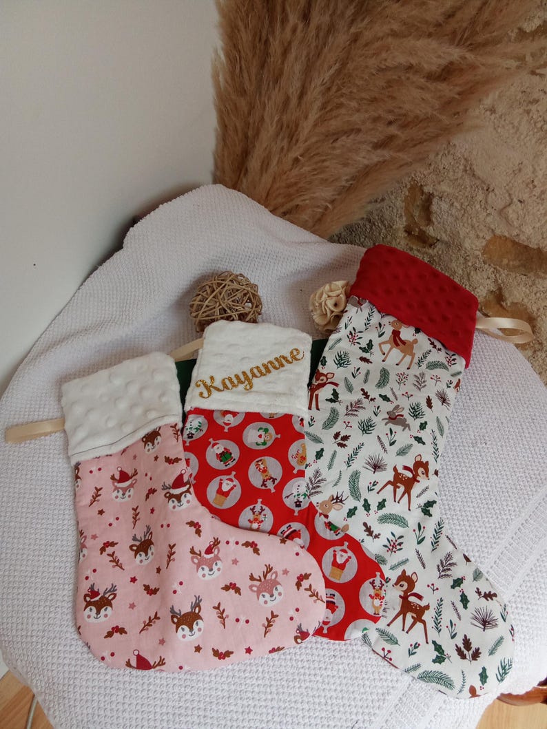K&ouml;nnte beinhalten: Drei Weihnachtsstr&uuml;mpfe mit einem Rentiermotiv. Der Strumpf links ist rosa mit wei&szlig;en Punkten, der mittlere Strumpf ist rot mit einem Weihnachtsmannmotiv und der rechte Strumpf ist wei&szlig; mit einem gr&uuml;nen und braunen Rentiermotiv. Der mittlere Strumpf tr&auml;gt den Namen "Kayanne" in goldener Stickerei.