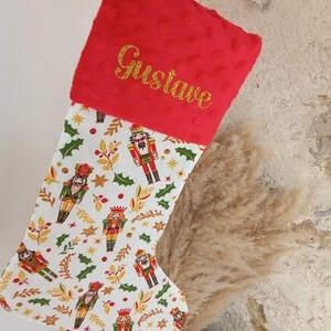 K&ouml;nnte beinhalten: Roter Weihnachtsstrumpf mit einem wei&szlig;en Stoff, der ein sich wiederholendes Muster aus Nussknackerfiguren, Stechpalmen und goldenen Sternen zeigt. Der Strumpf hat einen roten Pl&uuml;schbund mit dem Namen "Gustave", der in goldenen Glitzer gestickt ist.