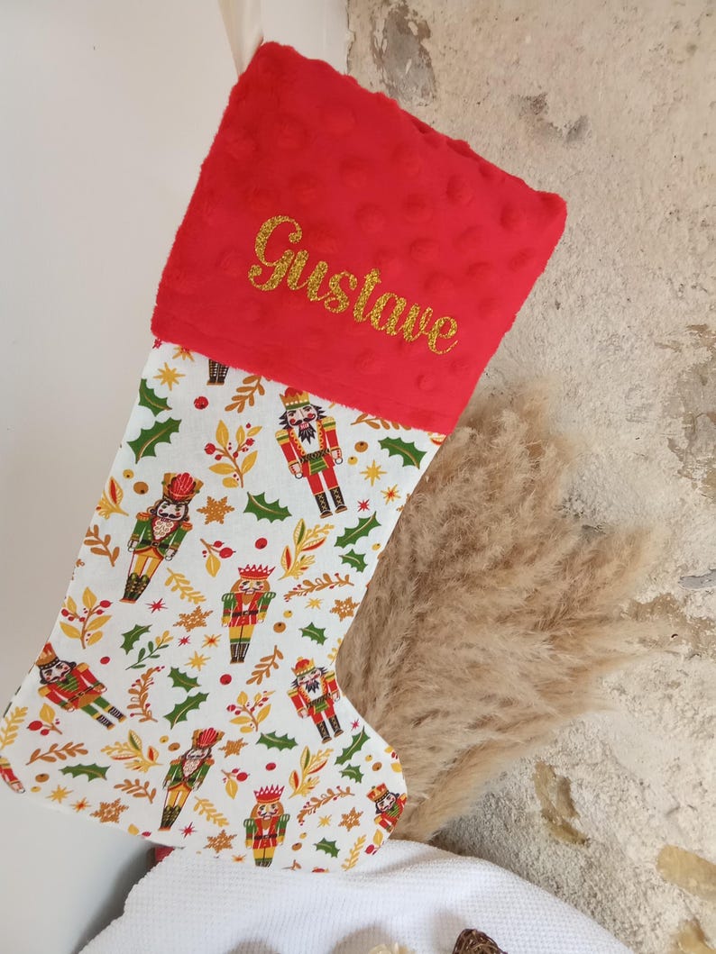 K&ouml;nnte beinhalten: Roter Weihnachtsstrumpf mit einem wei&szlig;en Stoff, der ein sich wiederholendes Muster aus Nussknackerfiguren, Stechpalmen und goldenen Sternen zeigt. Der Strumpf hat einen roten Pl&uuml;schbund mit dem Namen "Gustave", der in goldenen Glitzer gestickt ist.