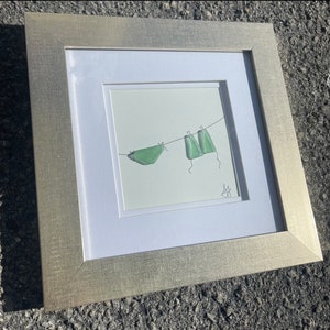 Puede incluir: Obra de arte enmarcada con una ilustración minimalista de trajes de baño verdes colgados de un tendedero. La obra de arte está sobre un fondo blanco, enmarcada en un marco plateado. La pieza tiene una estética sencilla pero elegante.