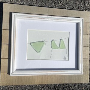 Puede incluir: Obra de arte enmarcada que muestra un bikini verde claro colgado de un tendedero. La obra está sobre un fondo blanco, enmarcada con un borde blanco y dentro de un marco de madera rústica. Diseño minimalista.