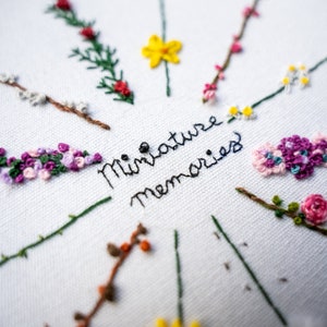 May include: White fabric with embroidered flowers in shades of pink, purple, and green. The text "Miniature Memories" is embroidered in black thread.