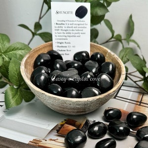 May include: A small wooden bowl filled with black shungite tumbled stones. The bowl is sitting on top of two books. The top book is titled "At Home With Old & New" by Joanna Gaines. The bottom book is titled "Shungite" by Mitchell Beazley. A small card with the text "Shungite" is visible in the background.