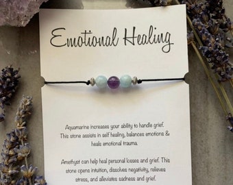 May include: A black string bracelet with aquamarine and amethyst beads. The bracelet is on a white card with the text "Emotional Healing" and information about the healing properties of the stones.
