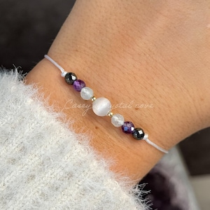 May include: A delicate bracelet strung on a white cord, featuring selenite, amethyst, aquamarine, and hematite beads. The bracelet has a central gold-colored bead. The text "Casey crystal cove" and the names of the stones are written on the image.