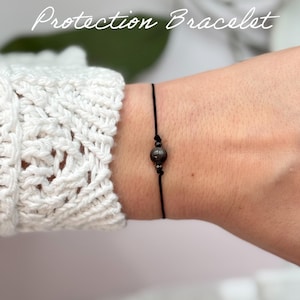 May include: A black string bracelet with a single dark grey bead. The bracelet is on a person's wrist. The text "Protection Bracelet" is above the bracelet and the text "Repels & blocks negative energy" is below the bracelet.