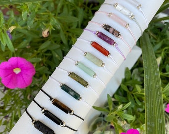 Könnte beinhalten: Eine Auswahl an bunten Edelstein-Armbändern mit silbernen Perlen und Schnur. Die Armbänder werden auf einem weißen Zylinder präsentiert.