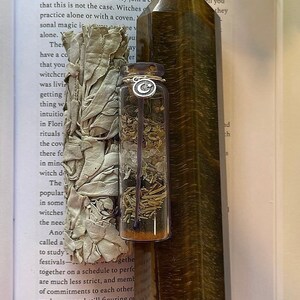 May include: A brown and gold tiger's eye crystal point, a small glass bottle filled with dried herbs, and a white sage smudge stick wrapped in white fabric. The items are arranged on a page of a book with text about witchcraft.