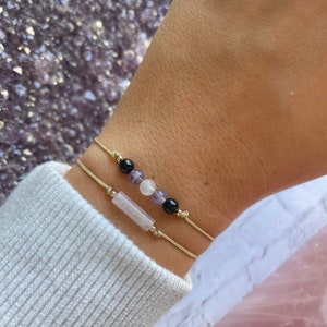 Puede incluir: Dos pulseras con hilo dorado y cuentas de piedras preciosas. Una pulsera tiene una piedra rectangular de color rosa claro y la otra tiene cuentas negras, blancas y moradas.