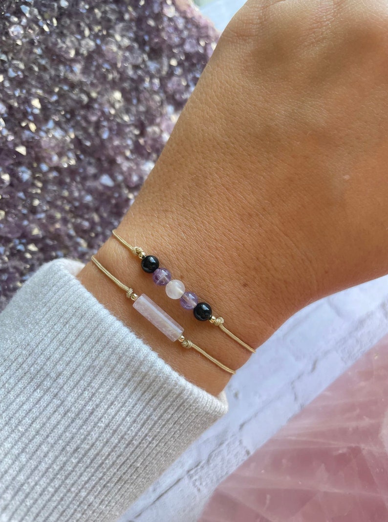Puede incluir: Dos pulseras con hilo dorado y cuentas de piedras preciosas. Una pulsera tiene una piedra rectangular de color rosa claro y la otra tiene cuentas negras, blancas y moradas.