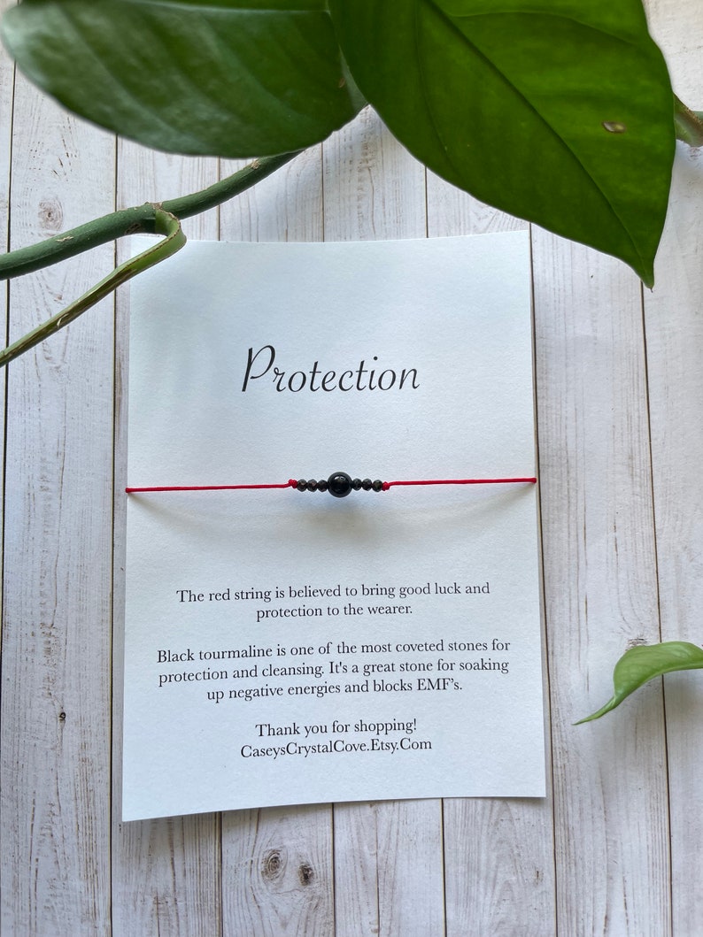 May include: Red string bracelet with black tourmaline beads. The card reads "Protection" and explains the benefits of the stones. "The red string is believed to bring good luck and protection to the wearer. Black tourmaline is one of the most coveted stones for protection and cleansing. It's a great stone for soaking up negative energies and blocks EMF's. Thank you for shopping! CaseysCrystalCove.Etsy.Com"