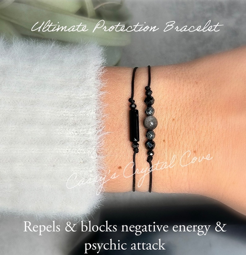 May include: Two black string bracelets with black and gray gemstone beads. The bracelets are on a person's wrist. The text "Ultimate Protection Bracelet" is at the top of the image. The text "Repels & blocks negative energy & psychic attack" is at the bottom of the image.