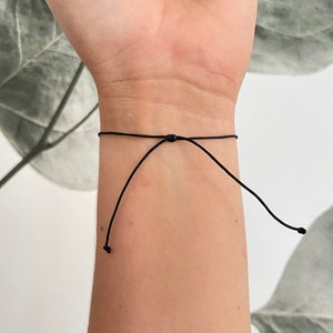 Puede incluir: Un brazalete ajustable simple hecho de cord&oacute;n delgado de color negro.