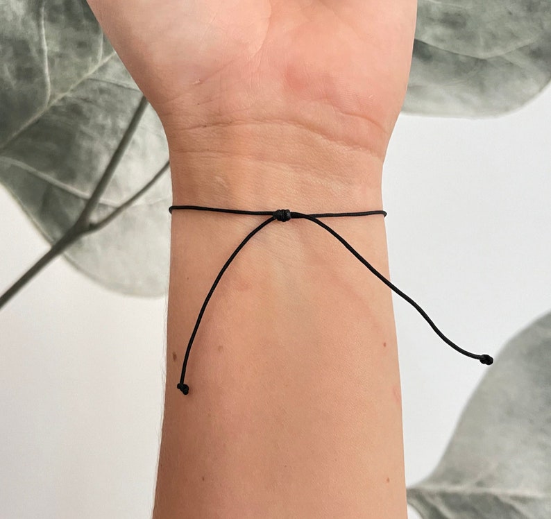 Puede incluir: Un brazalete ajustable simple hecho de cord&oacute;n delgado de color negro.