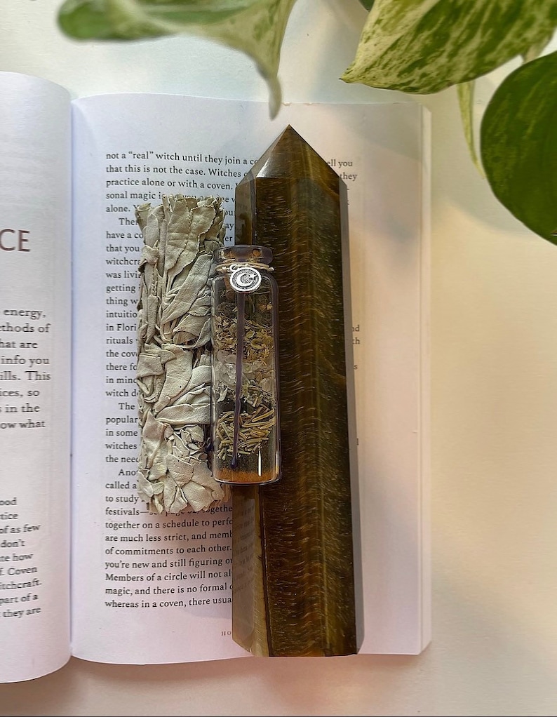 May include: A brown and gold tiger's eye crystal point, a small glass bottle filled with dried herbs, and a white sage smudge stick wrapped in white fabric. The items are arranged on a page of a book with text about witchcraft.