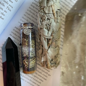 May include: A small glass bottle filled with herbs, crystals, and a silver charm. The bottle is sitting on a book with the word "Witchcraft" printed on the cover. The bottle is surrounded by a white sage smudge stick.