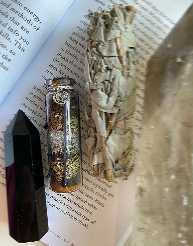 May include: A small glass bottle filled with herbs, crystals, and a silver charm. The bottle is sitting on a book with the word "Witchcraft" printed on the cover. The bottle is surrounded by a white sage smudge stick.