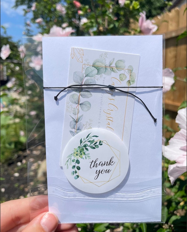 May include: A white card with a thank you sticker on it. The sticker has a gold geometric frame with green leaves and the words "thank you" in the center. The card is in a clear plastic bag.