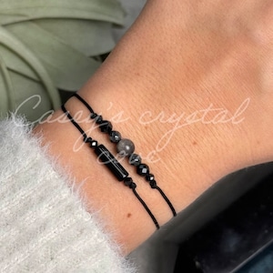 May include: Two black string bracelets with black beads and a grey stone bead. The bracelets are on a person's wrist.