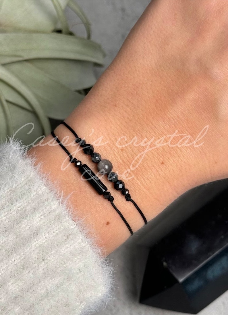 May include: Two black string bracelets with black beads and a grey stone bead. The bracelets are on a person's wrist.