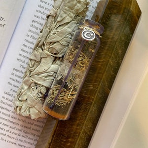 May include: A clear glass bottle filled with dried herbs and crystals, with a silver charm attached to the lid. The bottle is resting on a brown and gold tiger's eye crystal. A bundle of white sage is in the background.