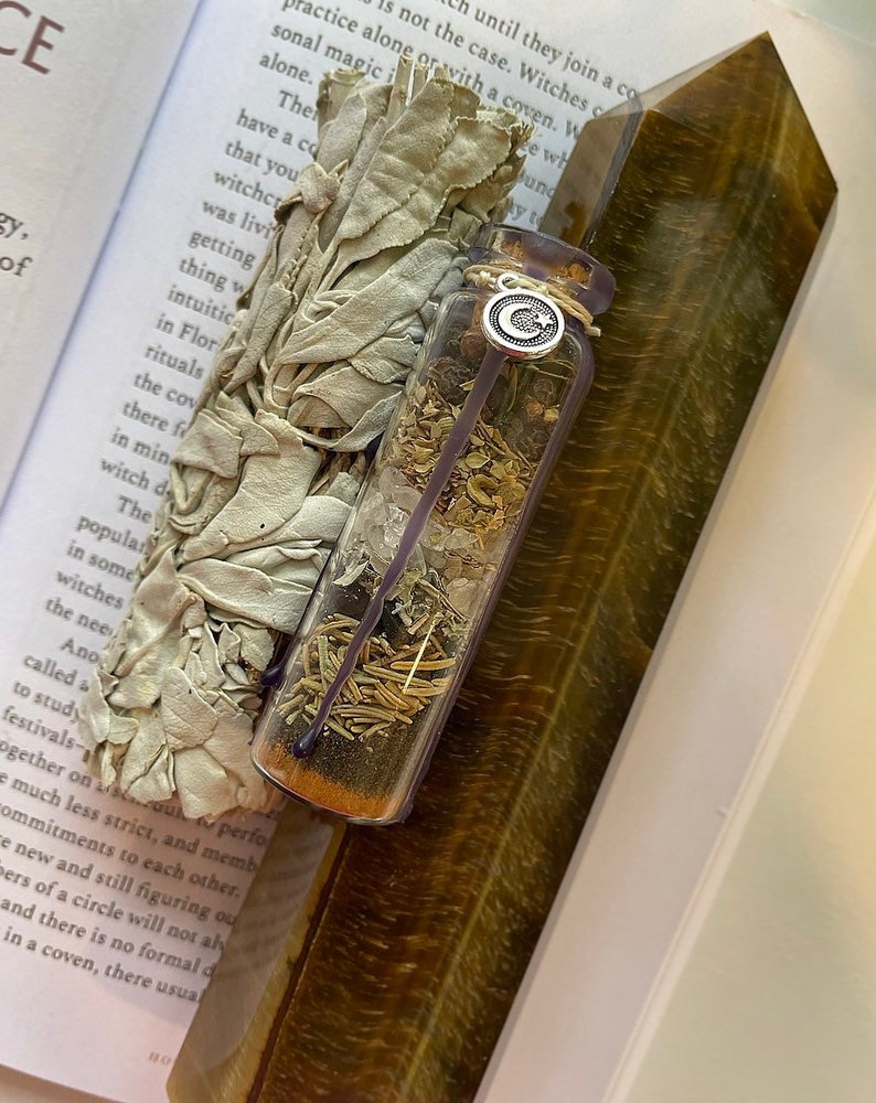 May include: A clear glass bottle filled with dried herbs and crystals, with a silver charm attached to the lid. The bottle is resting on a brown and gold tiger's eye crystal. A bundle of white sage is in the background.