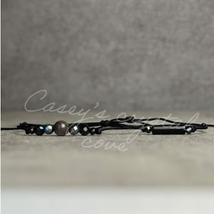 May include: Black beaded bracelet with a brown bead and a black rectangular bead. The bracelet is on a gray background.