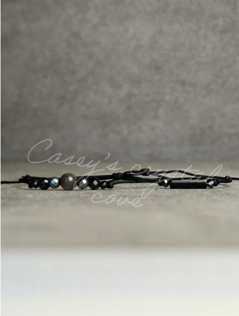 May include: Black beaded bracelet with a brown bead and a black rectangular bead. The bracelet is on a gray background.