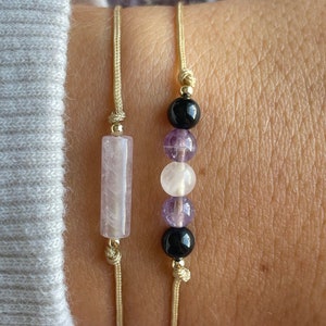 Puede incluir: Dos pulseras con cuentas de piedra natural. Una pulsera tiene una cuenta rectangular rosa y la otra tiene cuentas redondas negras, blancas y moradas.