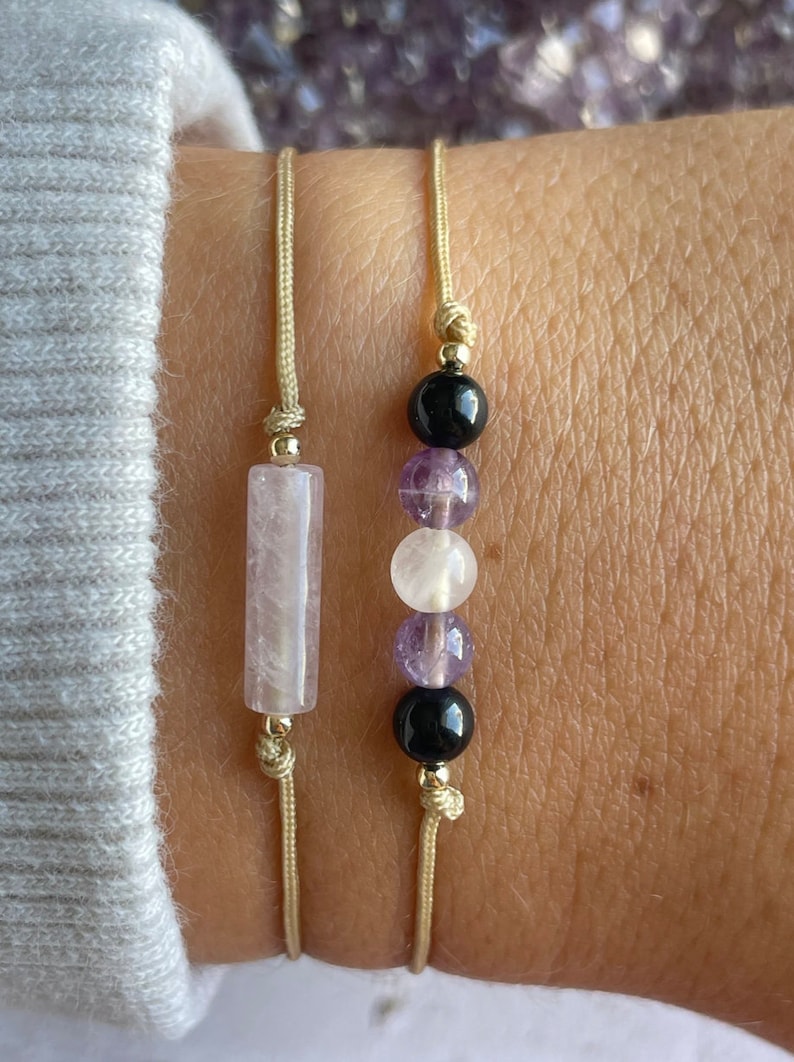 Puede incluir: Dos pulseras con cuentas de piedra natural. Una pulsera tiene una cuenta rectangular rosa y la otra tiene cuentas redondas negras, blancas y moradas.