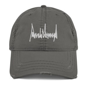 Peut inclure: Une casquette de baseball grise us&eacute;e avec une signature brod&eacute;e en blanc qui lit "Donald J. Trump".