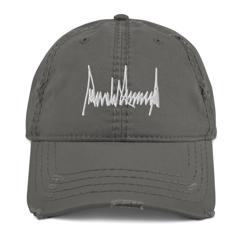 Peut inclure: Une casquette de baseball grise us&eacute;e avec une signature brod&eacute;e en blanc qui lit "Donald J. Trump".
