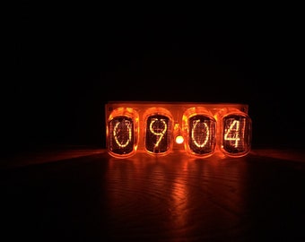 May include: A close-up of a four-digit Nixie tube clock displaying the time 09:04. The tubes are glowing orange and the numbers are clearly visible against the dark background.