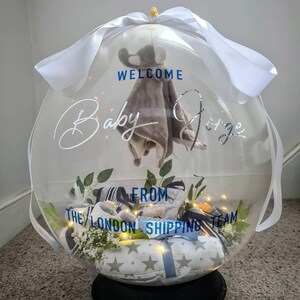 May include: A clear balloon filled with baby clothes and a stuffed animal. The balloon is decorated with a white ribbon and the text "Welcome Baby [name] from The London Shipping Team".
