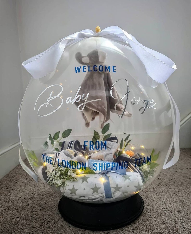 May include: A clear balloon filled with baby clothes and a stuffed animal. The balloon is decorated with a white ribbon and the text "Welcome Baby [name] from The London Shipping Team".