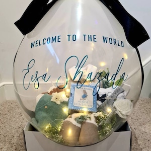 May include: A clear balloon filled with baby items and lights. The balloon is tied with a black ribbon and has the text "Welcome to the world Eesa Shazada" printed on it. The balloon is sitting on a white box with the same text printed on it.