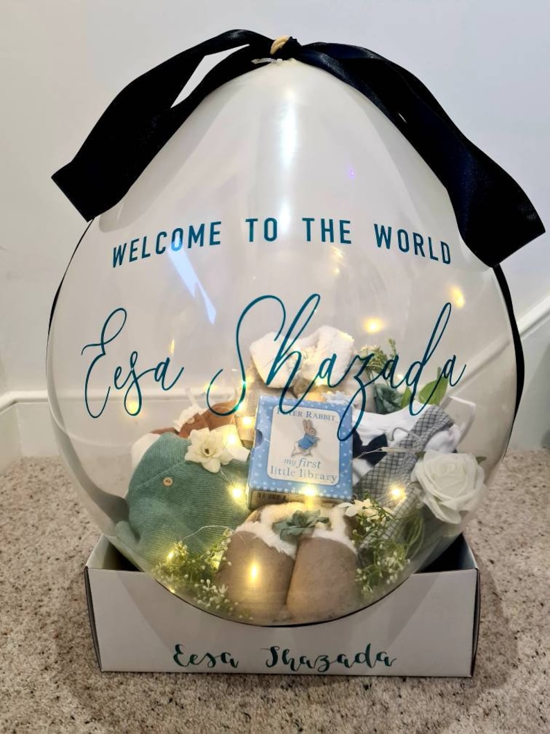 May include: A clear balloon filled with baby items and lights. The balloon is tied with a black ribbon and has the text "Welcome to the world Eesa Shazada" printed on it. The balloon is sitting on a white box with the same text printed on it.