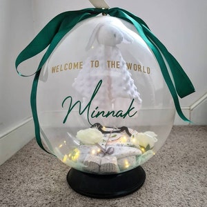 May include: A clear balloon filled with baby items, including a white stuffed bunny, a baby outfit, and a string of lights. The balloon is tied with a green ribbon and has the text "Welcome to the world. Minnak" printed on it.