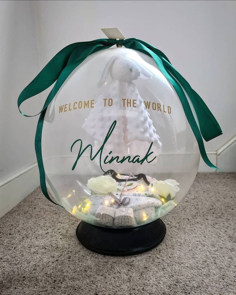 May include: A clear balloon filled with baby items, including a white stuffed bunny, a baby outfit, and a string of lights. The balloon is tied with a green ribbon and has the text "Welcome to the world. Minnak" printed on it.