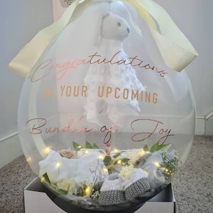 May include: A clear balloon filled with baby items, including a stuffed animal, a pair of booties, and a small blanket. The balloon is decorated with a gold ribbon and a white sticker that says "Congratulations on your upcoming bundle of joy".