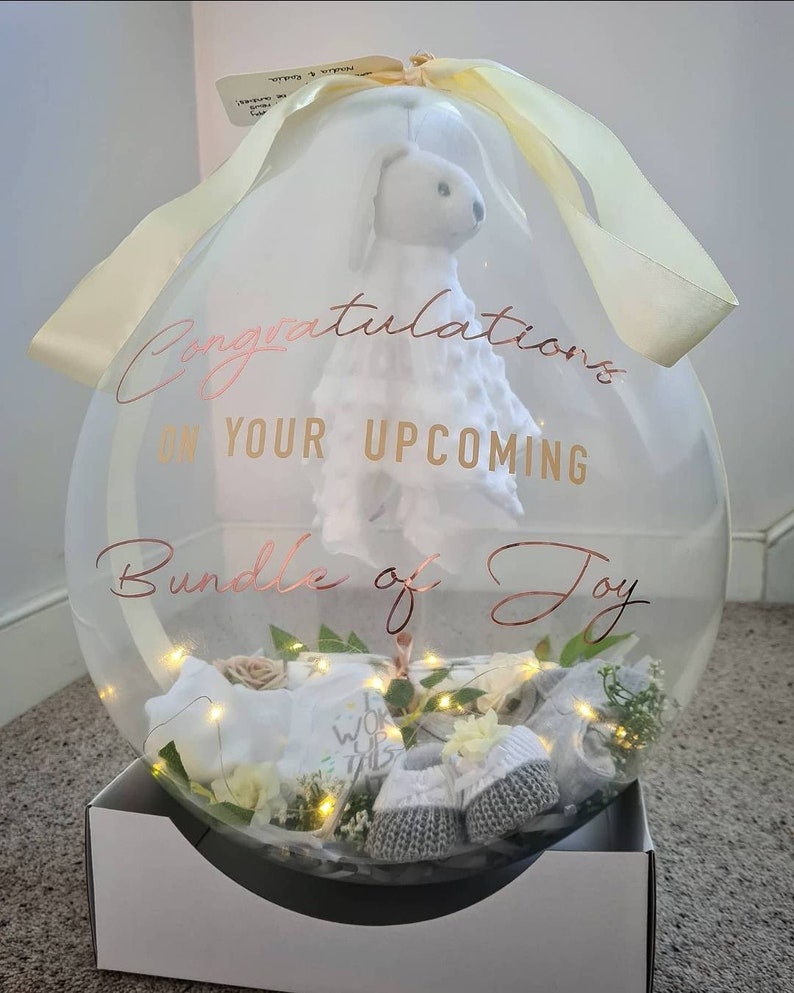 May include: A clear balloon filled with baby items, including a stuffed animal, a pair of booties, and a small blanket. The balloon is decorated with a gold ribbon and a white sticker that says "Congratulations on your upcoming bundle of joy".