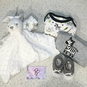 May include: A baby gift set with a white plush animal, a gray and white blanket, a white and gray patterned onesie, and a pair of gray knitted booties.