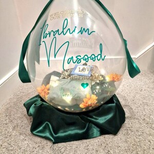 May include: A clear balloon with a green ribbon and a green satin base. The balloon has the text "Welcome to our Ummah Ibrahim Masood Love HuphOzara" written on it.